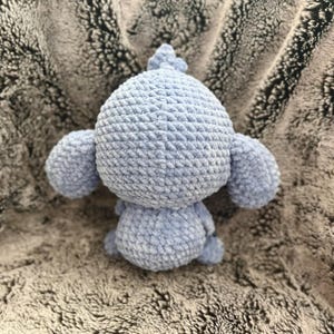 May include: A crocheted stuffed animal in the shape of a monkey. The monkey is light blue and has a textured surface. The monkey is sitting on a gray fuzzy surface.