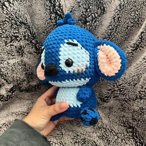 May include: A blue and pink crocheted plush toy resembling Stitch from the Disney movie Lilo & Stitch. The toy has a blue body with pink ears and a black nose.
