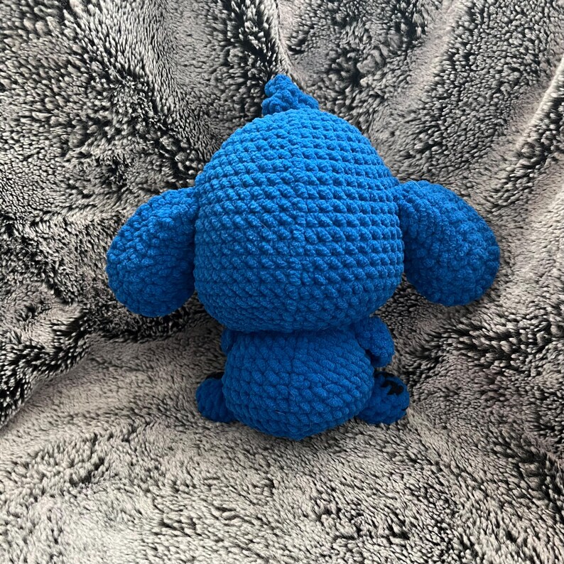May include: A blue crocheted stuffed animal with large ears. The animal is sitting on a fuzzy gray surface.