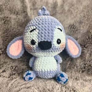 May include: A blue and white crocheted plush toy with large pink ears and blue feet. The toy has a black nose and large black eyes.