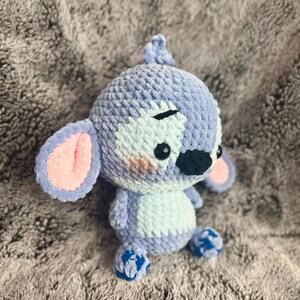 May include: A blue and white crocheted plush toy resembling a cartoon character with pink ears, black eyes, and a black nose. The toy is sitting on a gray fuzzy surface.