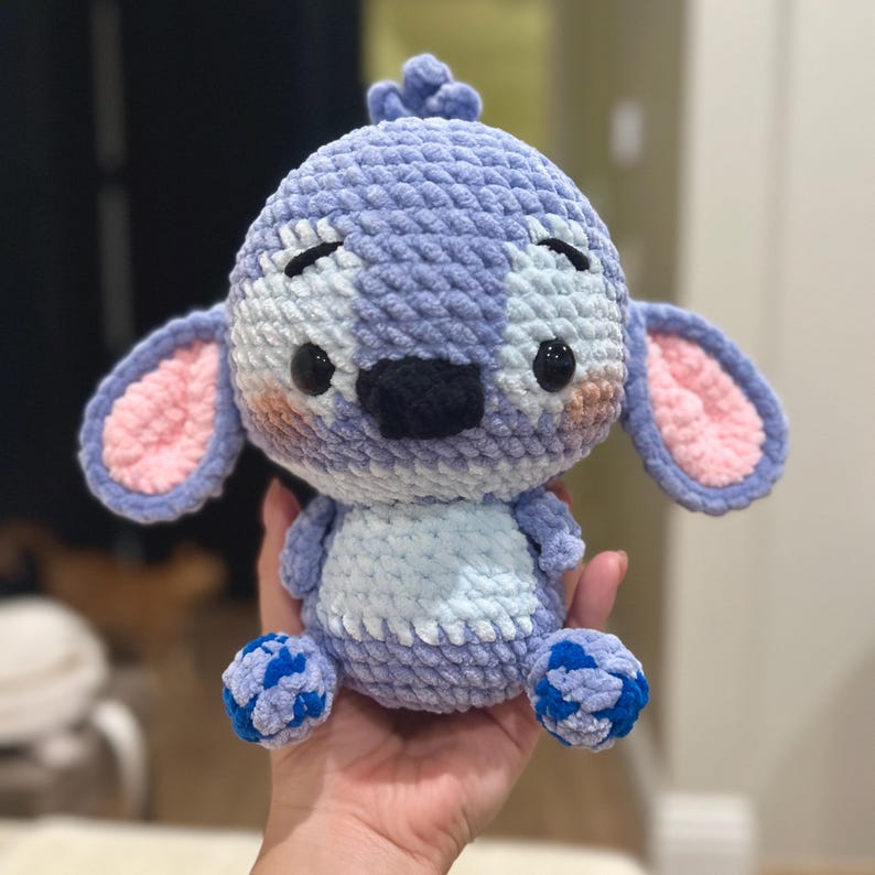 May include: A crocheted plush toy in the shape of a blue and white alien with pink ears and blue feet. The toy has a stitched-on black nose and black eyes.