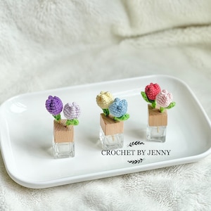 May include: Four small glass bottles with crocheted flower accents. The flowers are in shades of purple, yellow, blue, and pink. The bottles are sitting on a white plate. The text "CROCHET BY JENNY" is visible below the bottles.
