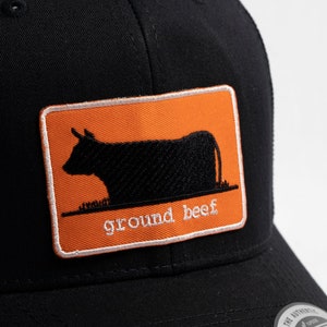 May include: Black trucker hat with an orange patch featuring a silhouette of a cow and the text "ground beef".