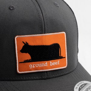 May include: A black and orange trucker hat with a patch featuring a silhouette of a cow and the text "ground beef".