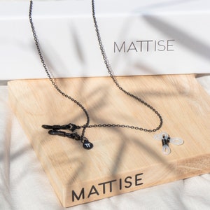 Op de afbeelding: Een zwarte metalen brillenketting met een delicaat kettingontwerp. De ketting heeft zwarte rubberen lussen voor het vastzetten van brillen en een kleine, ronde bedel. De ketting wordt weergegeven op een houten oppervlak met het woord "MATTISE".