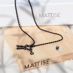 Op de afbeelding: Zwarte gevlochten brilketting met metalen uiteinden en een klein metalen bedeltje met de letter "T" erop. De ketting ligt op een houten oppervlak met het woord "MATTISE" erop gedrukt.