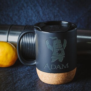 Peut inclure: Mug de voyage noir avec une anse et un couvercle noirs, orné d'un dessin de chevalier de dessin animé et du nom "ADAM". La base est en liège. Un citron jaune et un thermos argenté sont en arrière-plan.