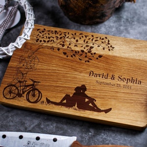 Könnte beinhalten: Personalisiertes Holzbrett zum Schneiden mit einem gravierten Design eines Paares, das unter einem Baum mit Fahrrädern und einem Vogelhaus sitzt. Das Brett ist mit dem Text "David & Sophia September 23, 2021" graviert.