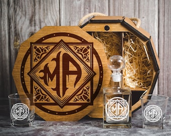 May include: A wooden box with a lid, containing a glass decanter and two glasses. The box is decorated with a brown and white monogram "MA" in a decorative frame. The decanter and glasses are also engraved with the same monogram.