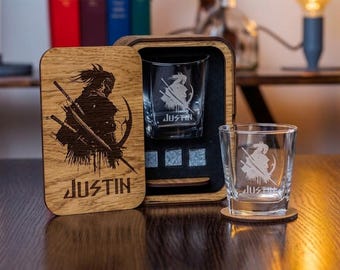 May include: A wooden box with a sliding lid featuring a laser-engraved samurai silhouette and the name JUSTIN. Inside the box are three grey stone cubes and a clear glass tumbler. A matching glass tumbler with the same engraving sits outside.