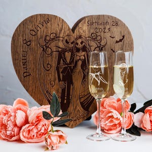 May include: A wooden heart-shaped box with a carved design of Jack Skellington and Sally from The Nightmare Before Christmas. The box is engraved with the names "Daniel & Olivia" and the date "September 28, 2023". Two champagne flutes are in front of the box, each engraved with a name.