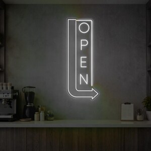 Könnte beinhalten: Ein weißes Neon-„OPEN“-Schild mit einem Pfeil, montiert an einer grauen Wand. Das Schild ist vertikal ausgerichtet, mit dem Wort „OPEN“ gestapelt. Der Pfeil zeigt nach rechts. Das Schild ist beleuchtet, und der Hintergrund ist neutral.