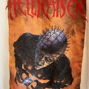 May include: A white t-shirt with a graphic print of a monster from the horror movie "Hellraiser". The monster is a pale, humanoid figure with a grid of nails on its head and a sharp, pointed hand. The text "Clive Barker's Hellraiser" is printed at the bottom of the graphic.