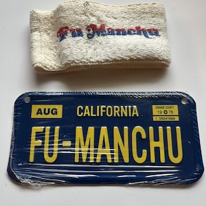May include: A white terry cloth wristband with red and blue embroidered text above a blue California license plate. The license plate is dark blue with yellow text that reads "FU-MANCHU". The license plate also has the text "AUG CALIFORNIA 19 76 ORANGE COUNTY J 06241999".