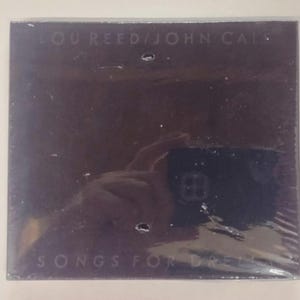 May include: Sealed CD case with the text "LOU REED / JOHN CALE" at the top and "SONGS FOR DRELLA" at the bottom. The case is dark with a reflective surface.