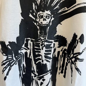 May include: Black and white graphic print of a skeleton with spiky hair and a wide open mouth. The skeleton is wearing a long robe and has its arms outstretched.