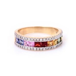 May include: A gold ring with a band of square-cut gemstones in a rainbow of colors, including blue, pink, red, and yellow. The ring is also adorned with a row of small, clear gemstones on the top and bottom edges.