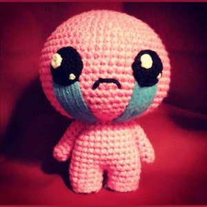 May include: A pink crocheted toy with a sad expression, large black eyes, and blue teardrop-shaped markings on its cheeks.