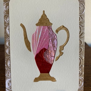 May include: A handmade greeting card with a gold-coloured teapot design and a pink and red interior. The teapot is set against a white background with a gold embossed border. The card is displayed on a clear stand.