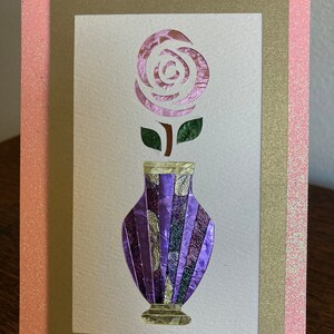 May include: A handmade greeting card featuring a purple vase with a rose. The rose is pink and the vase is purple with gold accents. The card has a pink glitter border and a gold inner border.