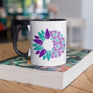 May include: White coffee mug with a black handle and black rim. The mug features a purple and teal sunflower design with a circle of purple ribbon symbols in the center.