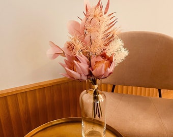 Op de afbeelding: Een boeket gedroogde roze en witte bloemen in een heldere glazen vaas met een bruine touw strik. De vaas staat op een gouden tafel.