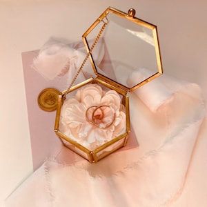 Pode incluir: Uma caixa de anel hexagonal dourada com tampa de vidro e uma flor branca dentro. Duas alianças de ouro estão aninhadas na flor.