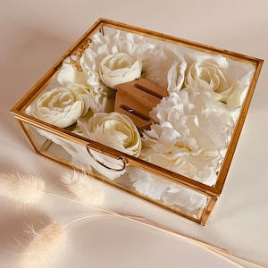 Pode incluir: Uma caixa de vidro com moldura dourada cheia de flores de seda brancas e uma peça decorativa de madeira. A caixa está sobre uma superfície branca.