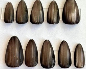 May include: Set of ten dark brown, oval-shaped press-on nails. Each nail features vertical, textured lines, creating a ribbed effect. The nails are arranged on a clear plastic strip against a white background.