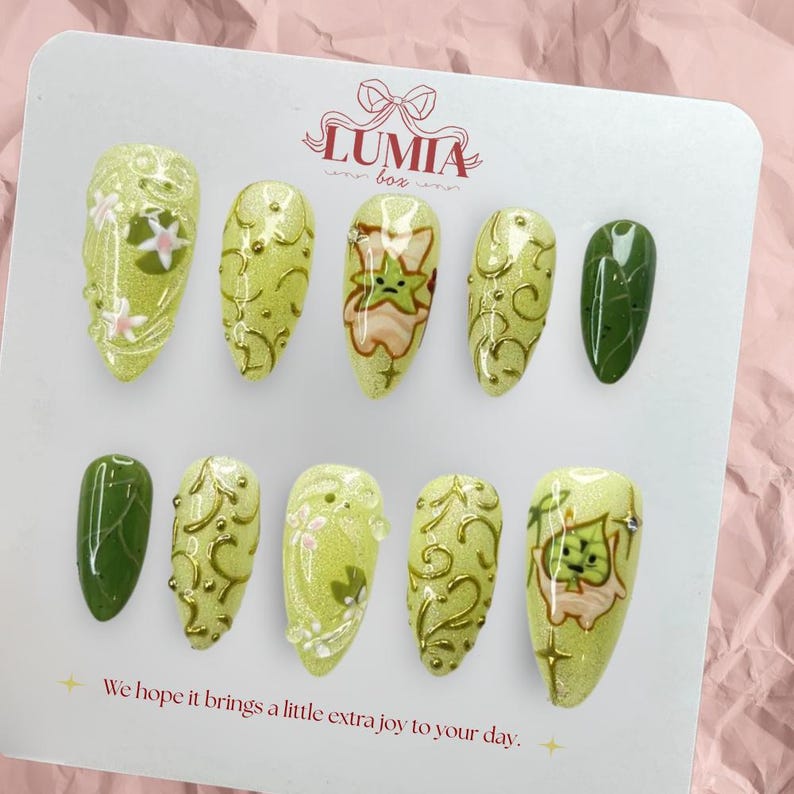 May include: A set of ten press-on nails in various designs. The nails are primarily light green with gold accents, floral patterns, and a cartoon character. The text "LUMIA box" is visible. The text "We hope it brings a little extra joy to your day." is also visible.