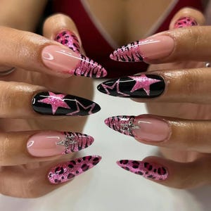 May include: A set of long, pointed acrylic nails. Some nails are black with pink star designs, others are pink with glitter leopard print, and some have a pink and silver design. The nails are on a person's fingers.