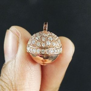 May include: A rose gold pendant with a round, dome-shaped design. The top half is covered in small, sparkling diamonds, while the bottom half is smooth. A small loop is at the top for a chain.