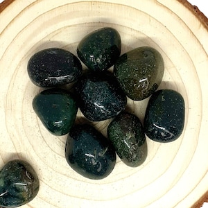 May include: A collection of eight smooth, dark green moss agate tumbled stones. The stones are arranged on a wooden surface.