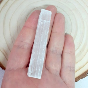 May include: A white selenite stick, also known as a selenite wand, is held in a hand. The stick is smooth and has a natural, striated texture.