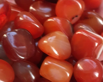 May include: A close-up of a collection of carnelian tumbled stones. The stones are a deep orange-red color and have a smooth, polished finish.