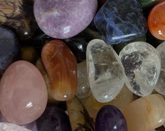 Puede incluir: Una colección de piedras preciosas pulidas de varios colores y formas. El surtido incluye piedras redondeadas en tonos de púrpura, rosa, naranja, azul y transparente. Algunas piedras tienen patrones y texturas visibles, creando una exhibición diversa.