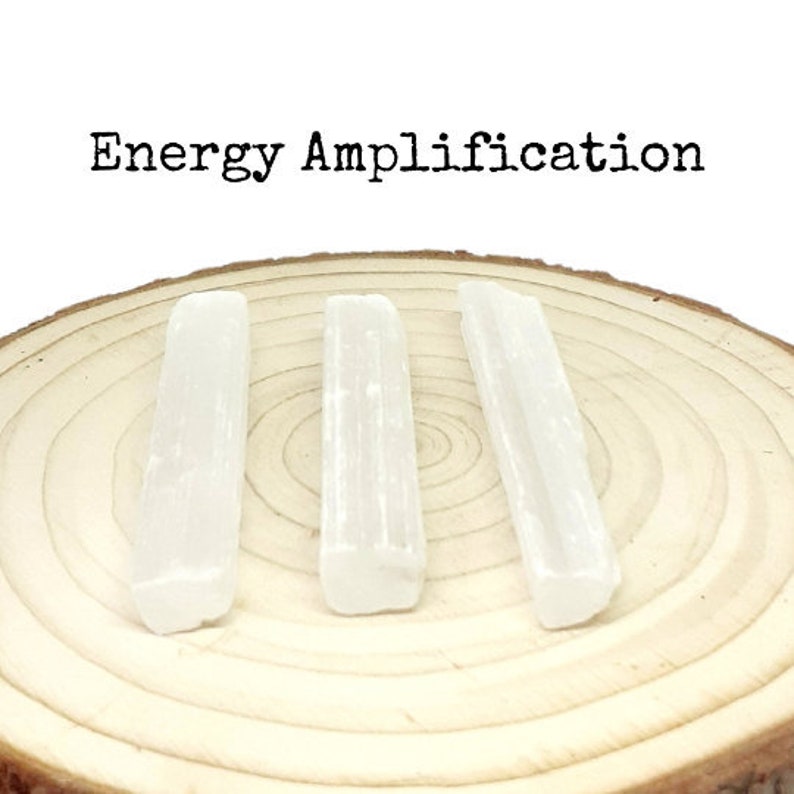 May include: Three white selenite sticks on a wooden surface. The sticks are smooth and have a natural, crystalline texture. The text "Energy Amplification" is written above the sticks.