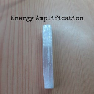 May include: A white selenite crystal wand, also known as a selenite stick, on a wooden surface. The text "Energy Amplification" is visible above the crystal.