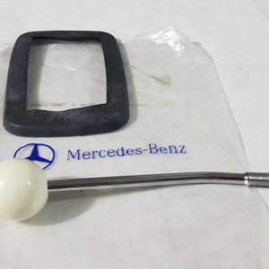 May include: A white plastic gear shift knob with a chrome gear shift lever and a black rubber gear shift boot. The gear shift lever is attached to the gear shift knob. The gear shift boot is in a plastic bag with the Mercedes-Benz logo.