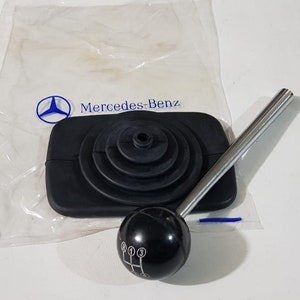 Puede incluir: Una funda de palanca de cambios de goma negra con una palanca de cambios de plata y una perilla de palanca de cambios negra con cinco marcas de cambios blancas. La funda de palanca de cambios está en una bolsa de plástico transparente con el logotipo de Mercedes-Benz.