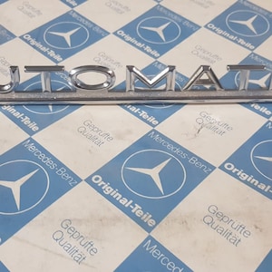 Puede incluir: Un emblema de coche de cromo plateado con la palabra "AUTOMATIC" en letras en relieve.