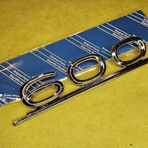May include: Chrome-finished "600" emblem with a horizontal mounting bar, resting on a blue and white package. The emblem is new and shiny, reflecting the light. The background is a solid yellow-green color.