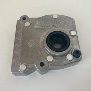 Op de afbeelding: Een zilverkleurig Mercedes-Benz auto-onderdeel. Het onderdeel heeft een rechthoekige vorm met afgeronde randen en een centrale cirkelvormige opening. De tekst "MERCEDES-BENZ 000 945 46 06" is zichtbaar. Er zijn ook verschillende kleine, cirkelvormige klinknagels aanwezig.