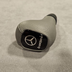 以下が含まれることがあります： メルセデス・ベンツのロゴと「Avantgarde」の文字が入った、黒いアクセントが付いたグレーの革製シフトノブ。シフトノブは丸みを帯びた形状で、上部は黒色です。