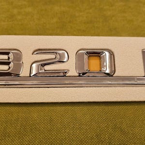 May include: A chrome car emblem displaying the numbers "320 E" on a rectangular backing. The emblem has a shiny, reflective finish, and the "0" features a yellow-orange square. This is likely a vehicle badge.