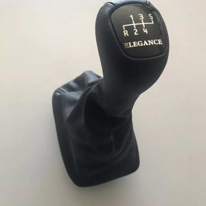 Könnte beinhalten: Schwarzer Leder-Schalthebelknauf mit einem silbernen Zahnradmuster und dem aufgedruckten Wort "Elegance".