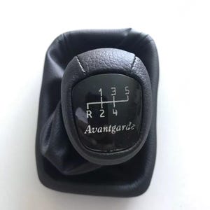 Può includere: Pomello del cambio in pelle nera con i numeri 1, 2, 3, 4 e 5, più R per la retromarcia. La parola "Avantgarde" è stampata sotto i numeri. Il pomello è circondato da un soffietto in pelle nera.