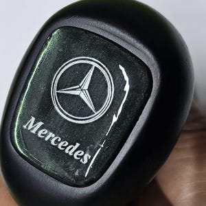 Peut inclure: Gros plan sur un pommeau de levier de vitesses Mercedes-Benz noir avec un logo Mercedes argenté et le mot "Mercedes" écrit en dessous.