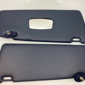Puede incluir: Dos viseras para el sol de coche de color azul oscuro con una superficie texturizada. Las viseras tienen un pequeño espejo rectangular en la parte superior y un clip en el lateral.